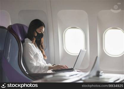 Asian young woman wearing protective mask using laptop sitting near windows at first class on airplane during flight,Traveling and Business during COVID-19 virus pandemic concept