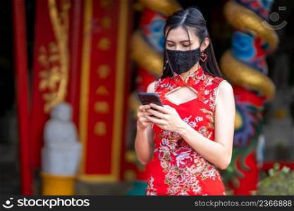 Asian young woman wearing red traditional Chinese cheongsam decoration and wear protective mask and smartphone for Chinese New Year Festival at shrine,Prevention of the spread of COVID-19 virus