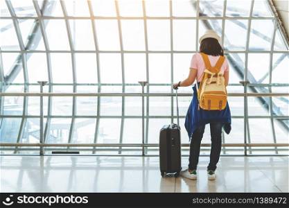 Asian young woman with suitcase and yellow backpack waiting for the flight at window of the airport. Rear view. Female is checking flight with smartphone. Copy space.