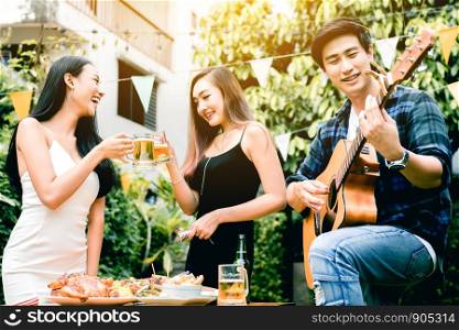 Asian young women enjoying toasting drinks party with guy playing guitar singing at home garden outdoors.