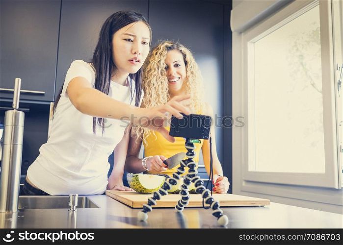 asiatic girl and caucasian girl making a video for a blog with fruit in the kitchen