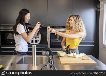 asiatic woman filming caucasian woman cutting fruit in a kitchen