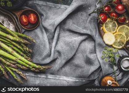 Asparagus cooking preparation with lemon and tomatoes on rustic napkin background, top view, frame. Dark style