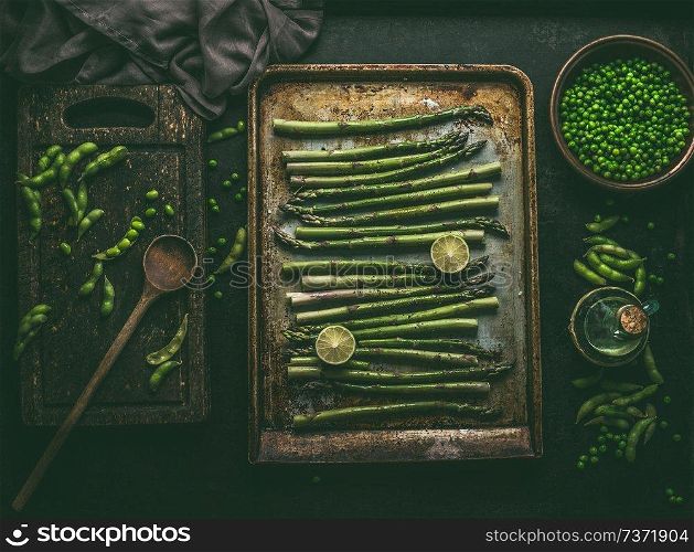Asparagus on baking tray with green ingredients : edamame soybeans, lime and green peas on dark rustic kitchen table background, top view.  Healthy vegetarian food