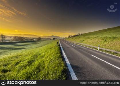 Asphalt road along the dike. Pasture on the background of Alps in Switzerland at sunset. Swiss landscape with meadows along the irrigation canal. . Asphalt road along the pasture