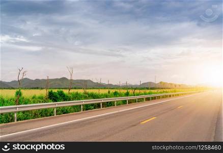 Asphalt road beside green grass field and the mountain with sunlight. Long distance journey with blue sky and white cloud. Country asphalt road. Road trip travel concept. Nature landscape.