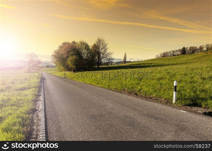 Asphalt road between pastures in Switzerland at sunrise. Swiss small town at the foot of mountains surrounded by meadows in the morning mist