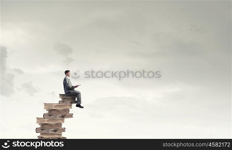 Aspiration for education. Young businessman sitting on pile of old book with one in hands