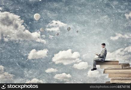 Aspiration for education. Young businessman sitting on pile of old book with one in hands