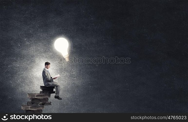 Aspiration for education. Young businessman sitting on pile of old books with one in hands