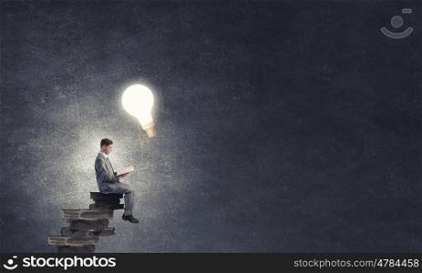 Aspiration for education. Young businessman sitting on pile of old books with one in hands