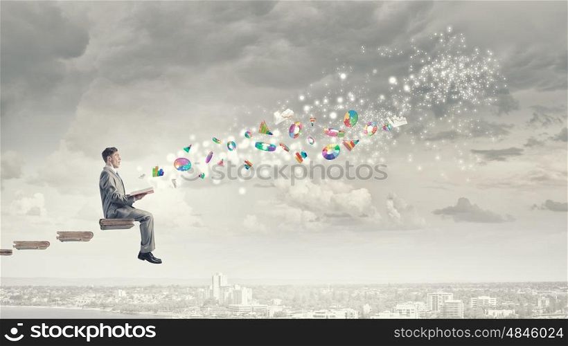 Aspiration for knowledge. Young businessman sitting on pile of books with one in hands and graphs flying in air