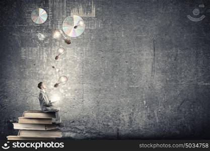 Aspiration for knowledge. Young businessman sitting on pile of books with one in hands and looking at graphs above