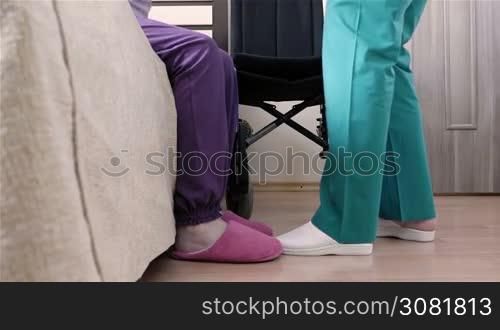 Assistant nurse helping senior disabled woman to put her in wheelchair at home or in nursing center. Slow motion