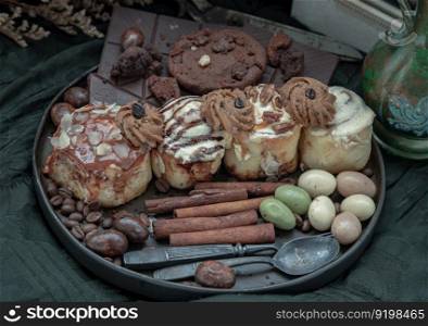 Assorted Cinnamon Rolls, Dark chocolate bars, cookies, pieces and balls served with black iron cutlery in black ceramic plate on dark background. Selective Focus.
