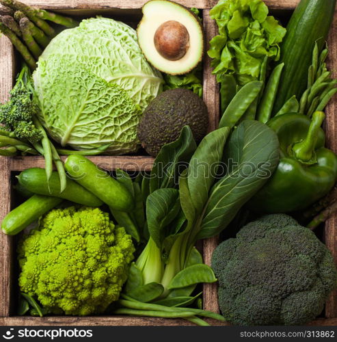 Assorted green toned raw organic vegetables in wooden box on dark stone background. Avocado, cabbage, cauliflower and cucumber with trimmed and mung beans and pepper and broccoli with sparagus tips.