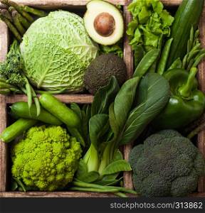 Assorted green toned raw organic vegetables in wooden box on dark stone background. Avocado, cabbage, cauliflower and cucumber with trimmed and mung beans and pepper and broccoli with sparagus tips.