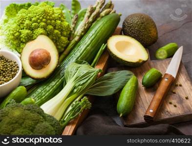 Assorted green toned raw organic vegetables in wooden box on dark stone background. Avocado, cabbage, cauliflower and cucumber with trimmed beans and chopping board with knife.