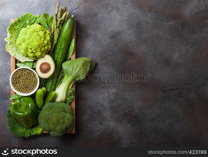 Assorted green toned raw organic vegetables in wooden box on dark stone background. Avocado, cabbage, cauliflower and cucumber with trimmed and mung beans and pepper and broccoli with sparagus tips.