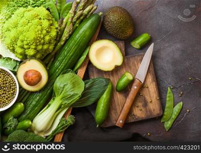 Assorted green toned raw organic vegetables in wooden box on dark stone background. Avocado, cabbage, cauliflower and cucumber with trimmed beans and chopping board with knife.