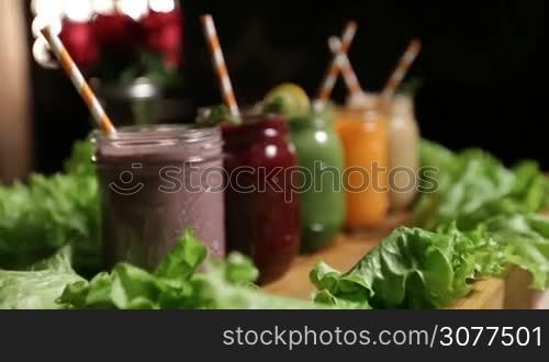 Assortment of fruit and vegetable smoothies in mason jars with striped straws, mint leaves and lemon slices on wooden tray decorated with green salad circle closeup. Healthy vegetarian diet for weight loss and detox.