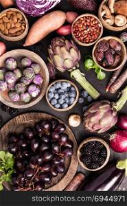 Assortment raw organic of purple ingredients: eggplants, artichokes, potatoes, onions, berries, nuts, carrots, brussel sprouts, grapes over dark background. Top view with space. Food frame