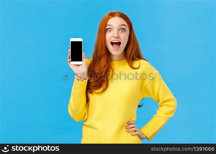 Astonished and impressed, excited redhead female in yellow sweater introduce new app, showing smartphone display, smiling fascinated open mouth amused, look camera, blue background.. Astonished and impressed, excited redhead female in yellow sweater introduce new app, showing smartphone display, smiling fascinated open mouth amused, look camera, blue background