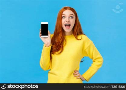 Astonished and impressed, excited redhead female in yellow sweater introduce new app, showing smartphone display, smiling fascinated open mouth amused, look camera, blue background.. Astonished and impressed, excited redhead female in yellow sweater introduce new app, showing smartphone display, smiling fascinated open mouth amused, look camera, blue background