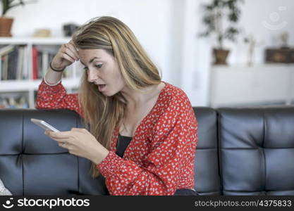 Astonished blond woman using a mobile phone while sitting on a leather sofa