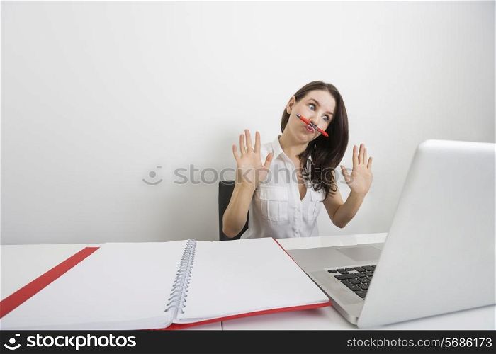 Astonished businesswoman looking at laptop while holding pen under nose in office