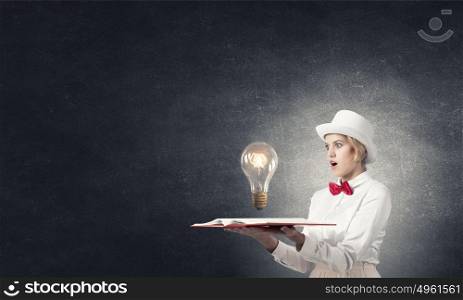 Astonished woman looking in book. Young woman in white hat with opened book in hands and glass light bulb on pages