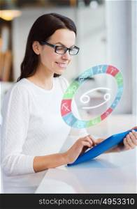 astrology, horoscope, people and technology concept - smiling young woman in eyeglasses with tablet pc computer and cancer zodiac sign at cafe or office. woman with tablet pc and zodiac signs at cafe