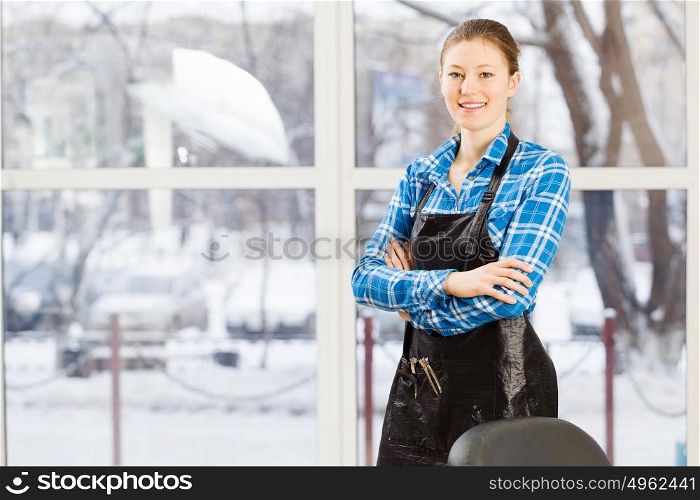 At beauty salon. Young woman hairdresser in apron standing near window