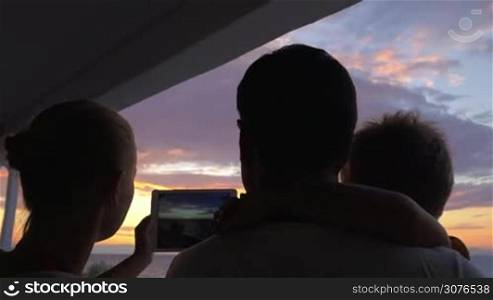 At sunset father stands with son in his arms, and mother is photographed on tablet of sunset and sea in city Perea, Greece