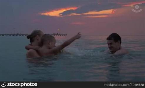 At sunset mother holding her son in her arms and swim in sea of city Perea, Greece, then swims up to them their dad and they begin to splash in the water. Happy family having fun