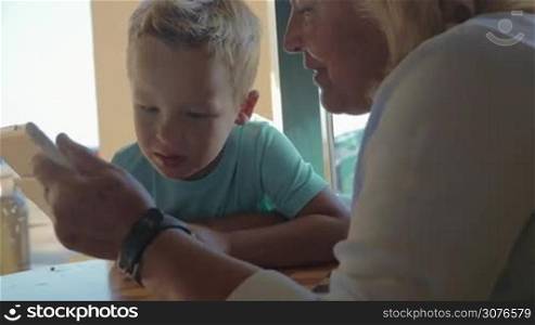 At table at cafe in city of Perea, Greece sits a grandmother with her grandson and teaches him how use tablet