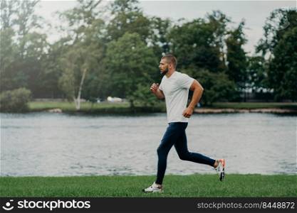 Athlete bearded man runs along beautiful landscape, demonstrates endurance, dressed in active wear, goes jogging near river, breathes fresh air. Sport, exercising, fitness and workout concept