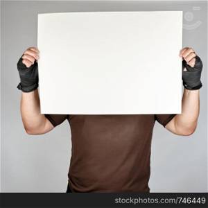 athlete in brown clothes holds a large blank white sheet of paper, his hands are wrapped in a black sports textile bandage, copy space