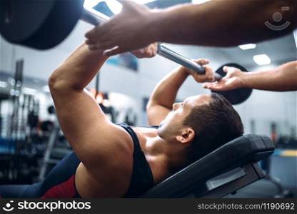 Athlete lies on bench and doing exercise with barbell under instructor control, motivation method, gym interior on background. Weightlifting workout in sport or fitness club. Athlete lies on bench, exercise with barbell