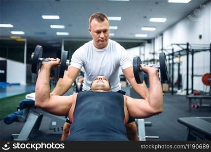 Athlete lies on bench and doing exercise with dumbbell under instructor control, motivation method, gym interior on background. Weightlifting workout in sport or fitness club. Athlete doing exercise under instructor control