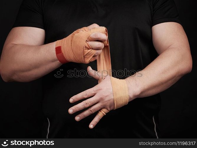 athlete stand in black clothes and wrap his hands in red textile elastic orange bandage before training, black background, muscular body