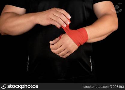 athlete stand in black clothes and wrap his hands in red textile elastic bandage before training, black background