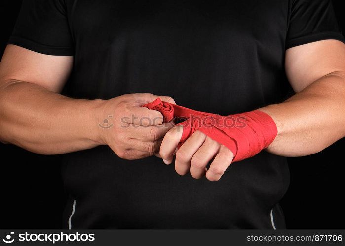 athlete stand in black clothes and wrap his hands in red textile elastic bandage before training, black background