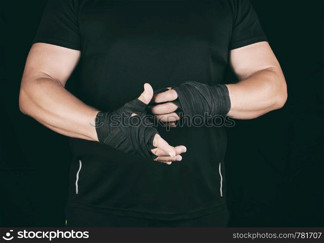 athlete stand in black clothes and wrap his hands in textile elastic bandage before training, dark background