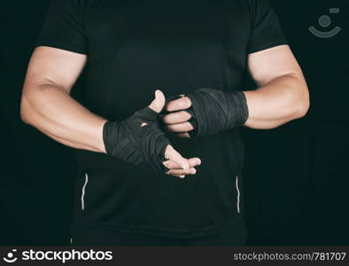 athlete stand in black clothes and wrap his hands in textile elastic bandage before training, dark background
