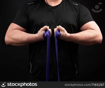 athlete with a muscular body in black clothes is doing physical exercises with blue rubber, low key
