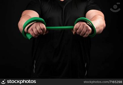 athlete with a muscular body in black clothes is doing physical exercises with green rubber, low key