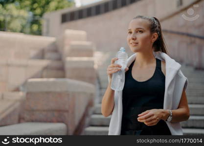 Athlete woman with pony tail feels thirsty after cardio training drinks water from bottle holds refreshig drink stays hydrated poses outdoors dressed in active wear concentrated into distance