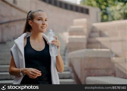Athlete woman with pony tail feels thirsty after cardio training drinks water from bottle holds refreshig drink stays hydrated poses outdoors dressed in active wear concentrated into distance