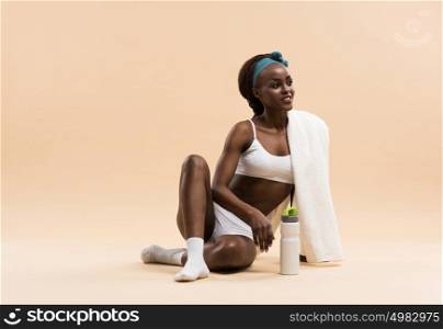 Athletic african girl with a bottle of water and towel sitting on beige studio background